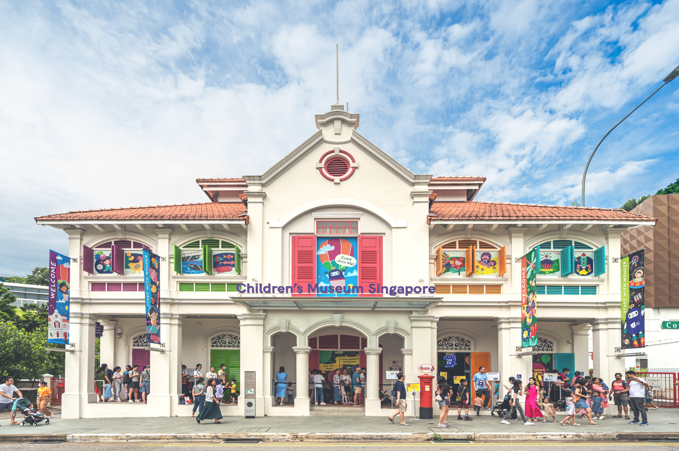 Children's Museum Singapore. A white building with colorful window shutters. People are gathered outside. Blue sky and clouds overhead.